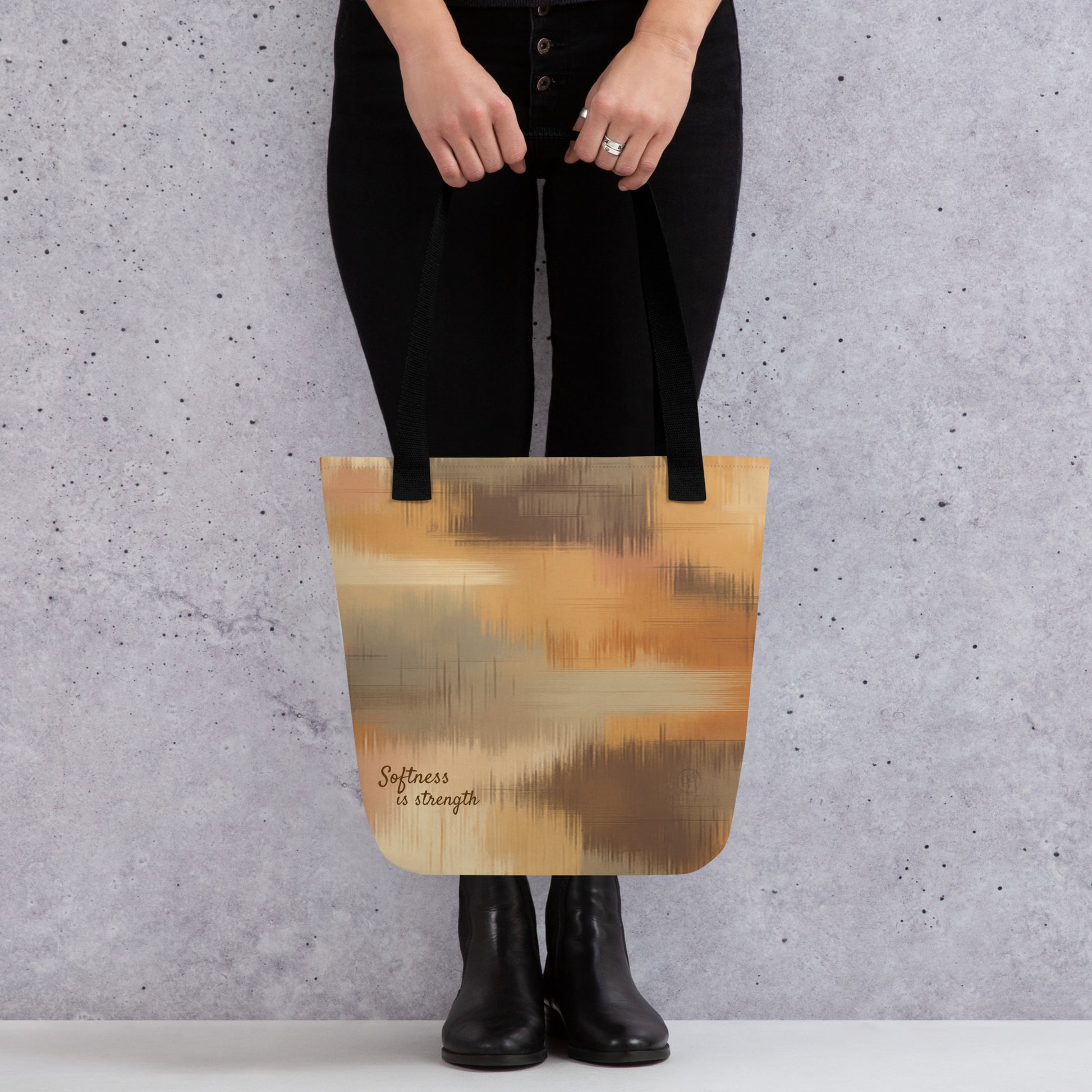 Person holding a beige and brown patterned tote bag with black handles and the phrase “Softness is strength,” standing against a grey wall.