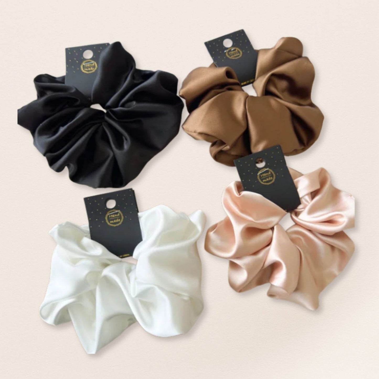 Four satin scrunchies in black, brown, white, and beige on a light beige background.