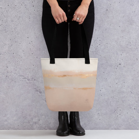 Person holding a beige and blush tote bag with gold brushstroke design and black straps, standing against a grey wall background.