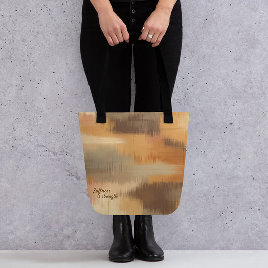 Person holding a beige and brown patterned tote bag with black handles and the phrase “Softness is strength,” standing against a grey wall.