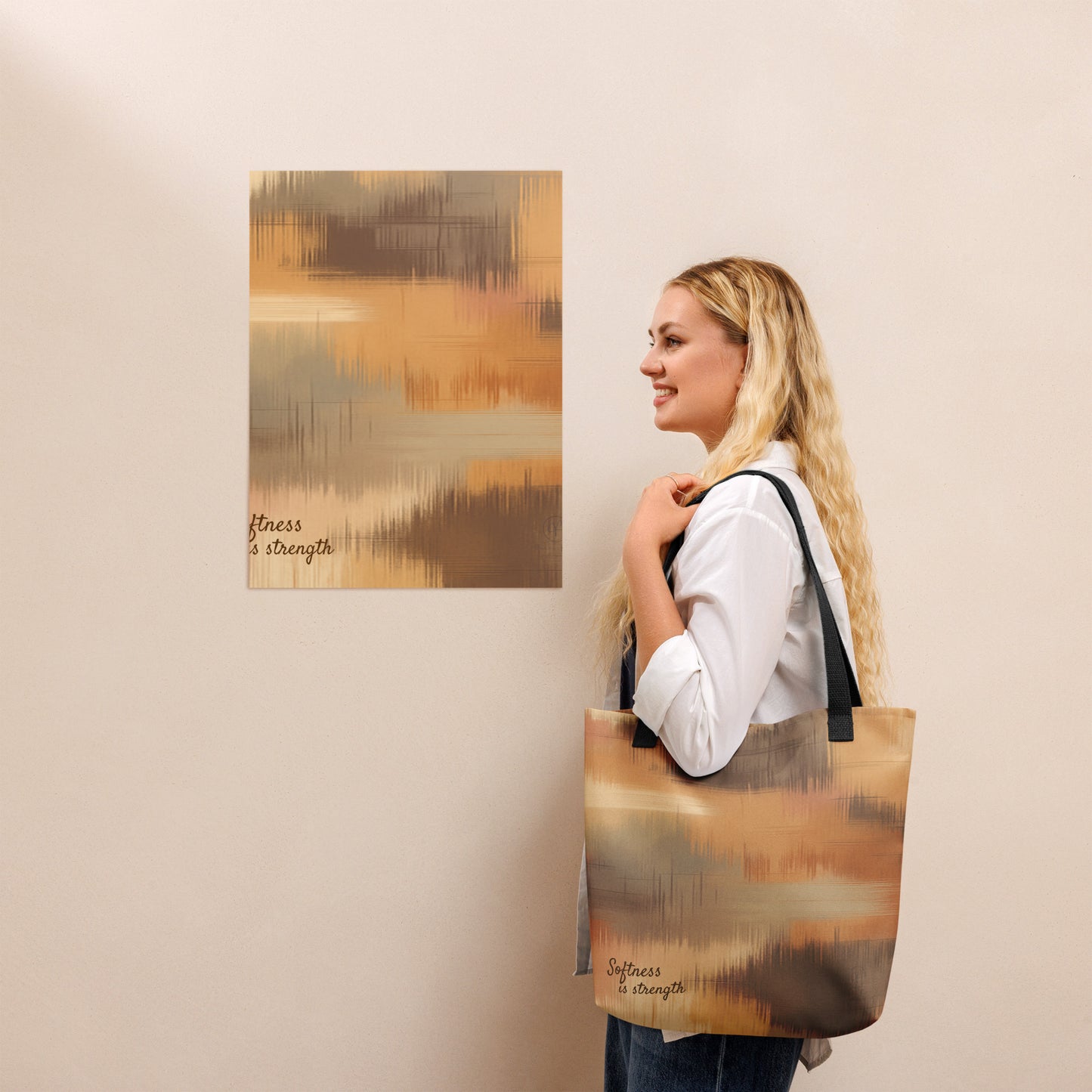 Person holding a beige and brown patterned tote bag with black handles, standing beside a matching abstract wall print with the phrase “Softness is strength.”