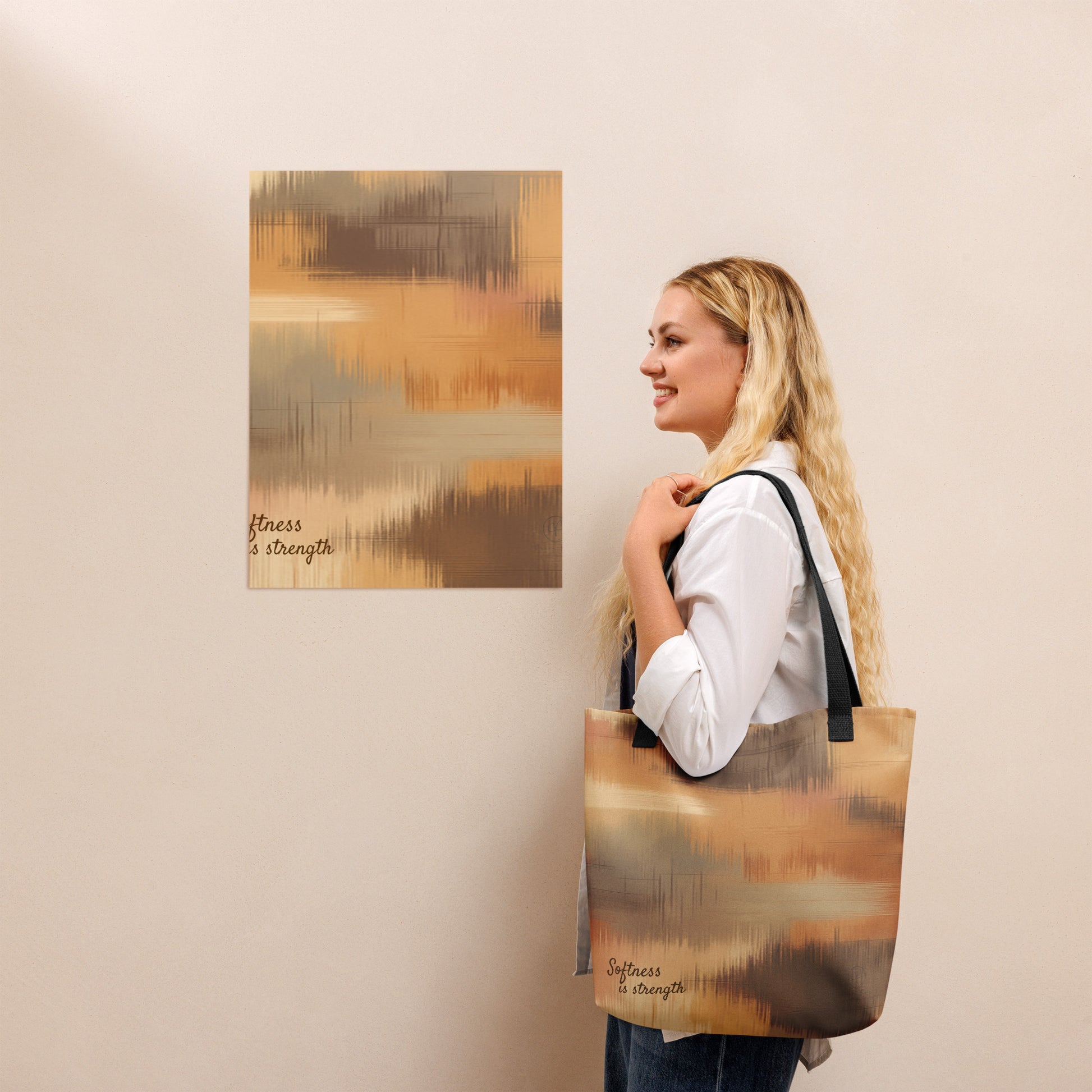 Person holding a beige and brown patterned tote bag with black handles, standing beside a matching abstract wall print with the phrase “Softness is strength.”